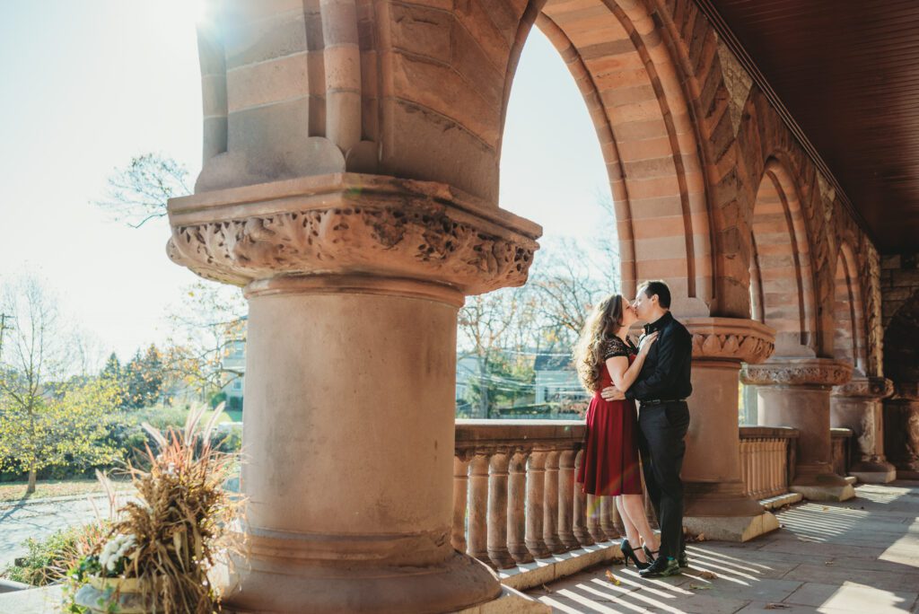 A romantic engagement session at the Queset Garden located at the Ames Public Library in Easton, Massachusetts. The couple is seen embracing amidst the garden's lush greenery and beautiful landscape, with the historic library building in the background, captured by Sarah Murray Photography.